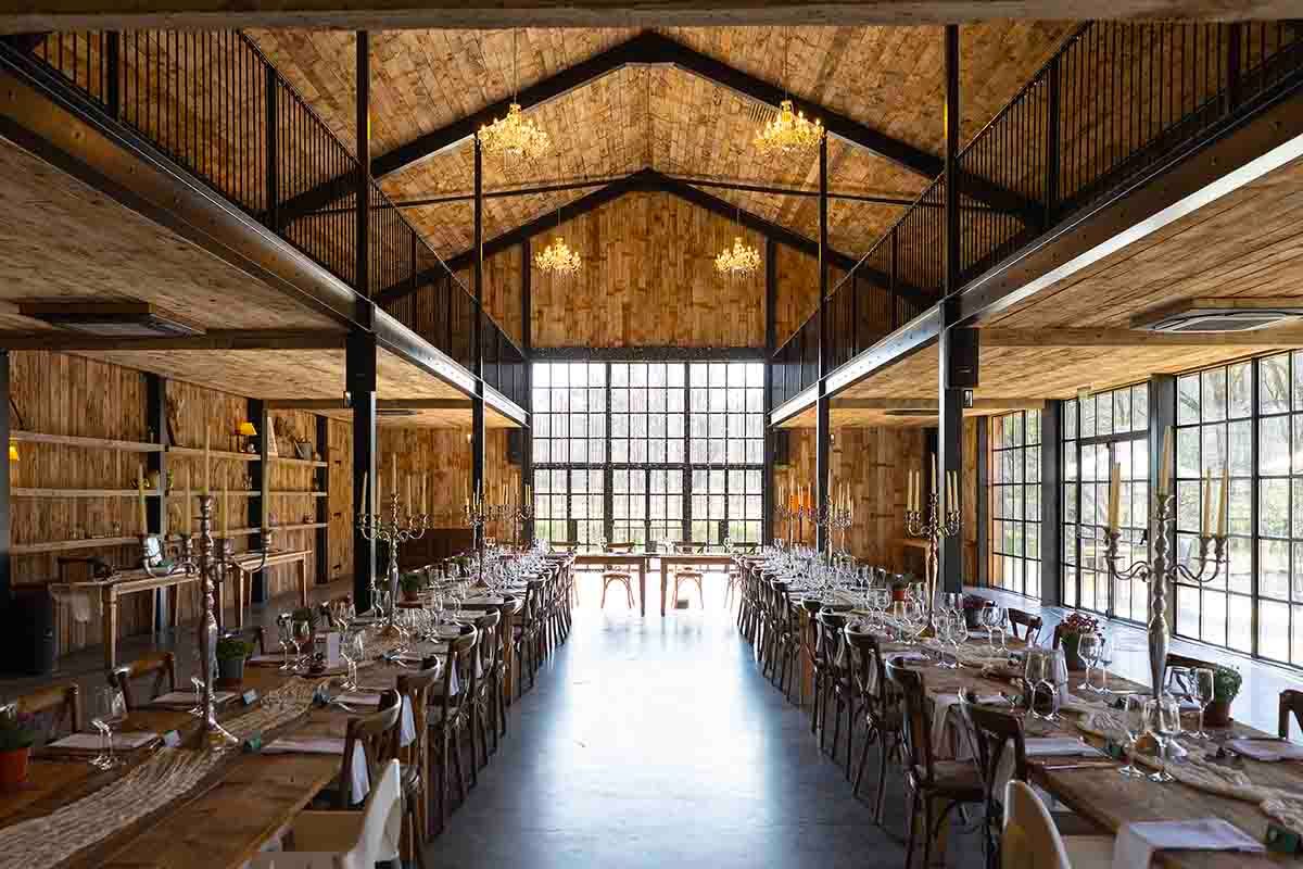 Rustic barn wedding venue with long dining tables, wooden walls, vaulted ceiling, and large windows bathed in warm light.
