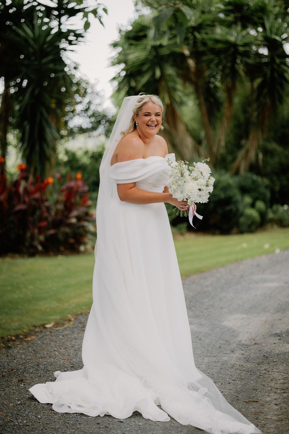 A bride in a flowing white wedding gown poses on a gravel path surrounded by lush tropical greenery.