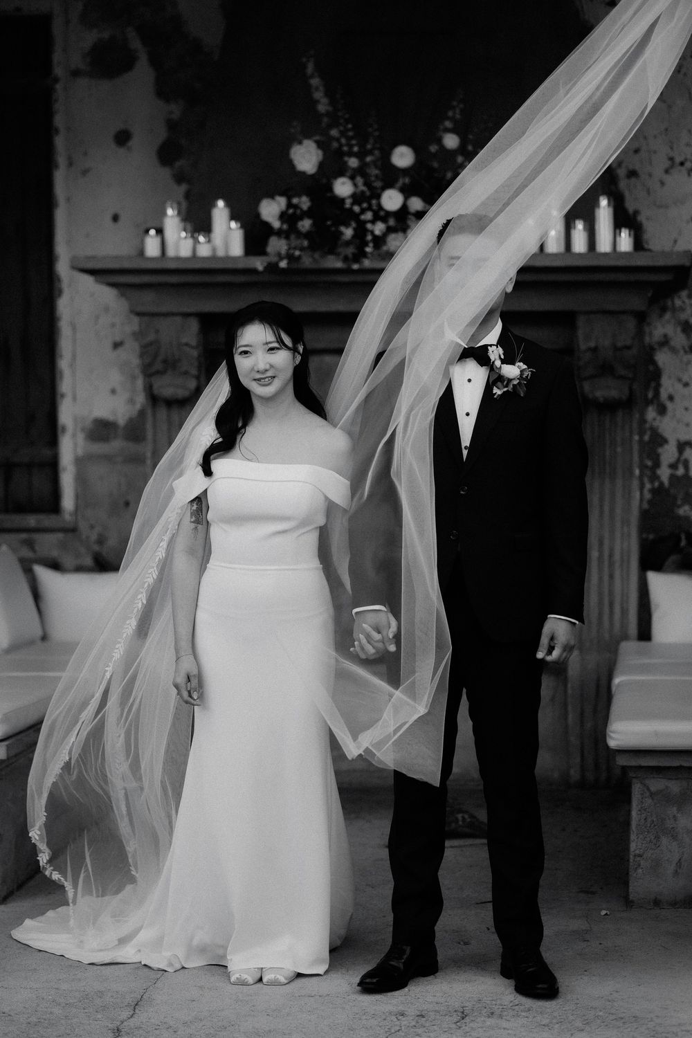A black and white wedding photo showing a couple with a flowing veil caught in the wind against a rustic backdrop.