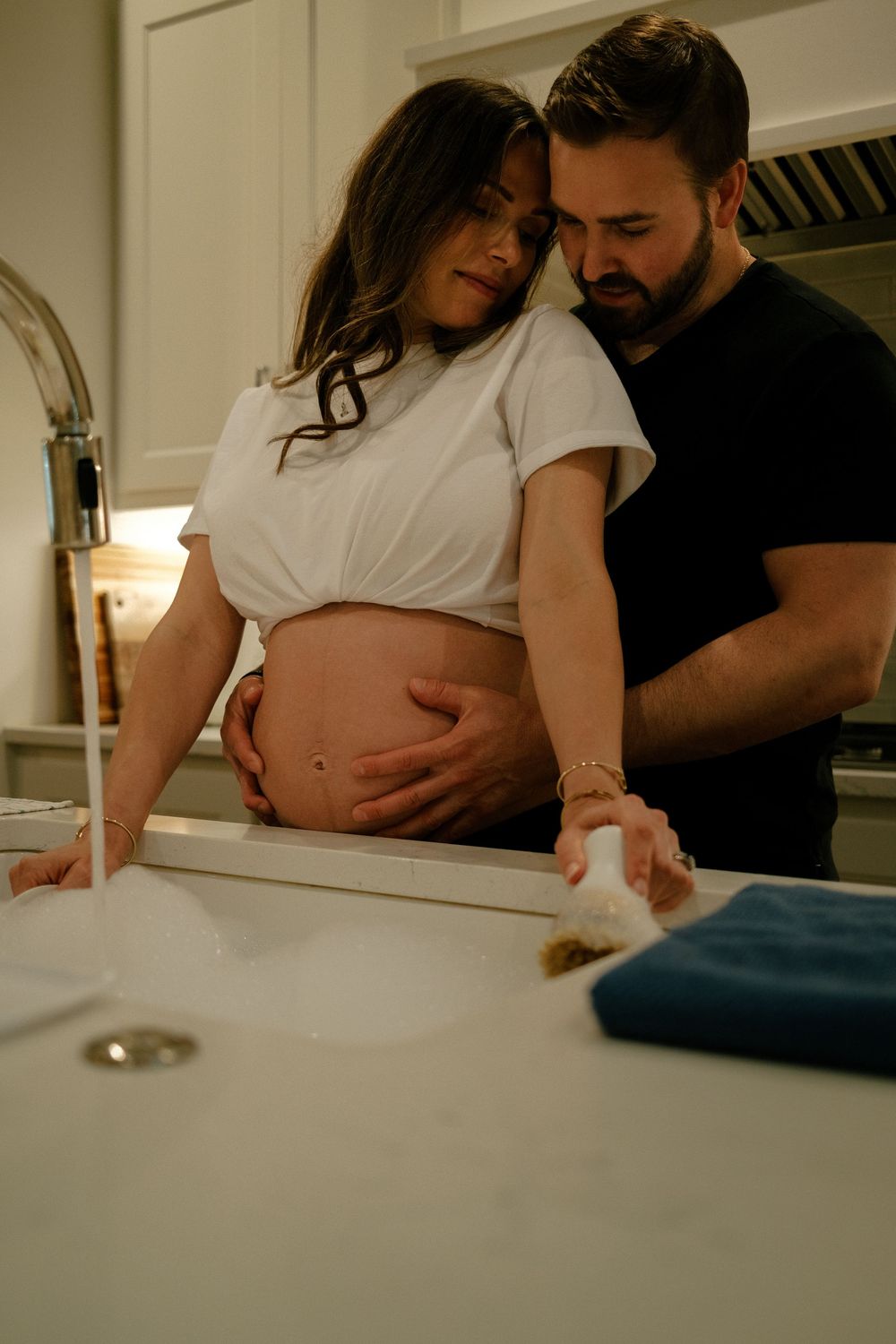 A couple shares a tender moment in the kitchen, with the man embracing his pregnant partner from behind.