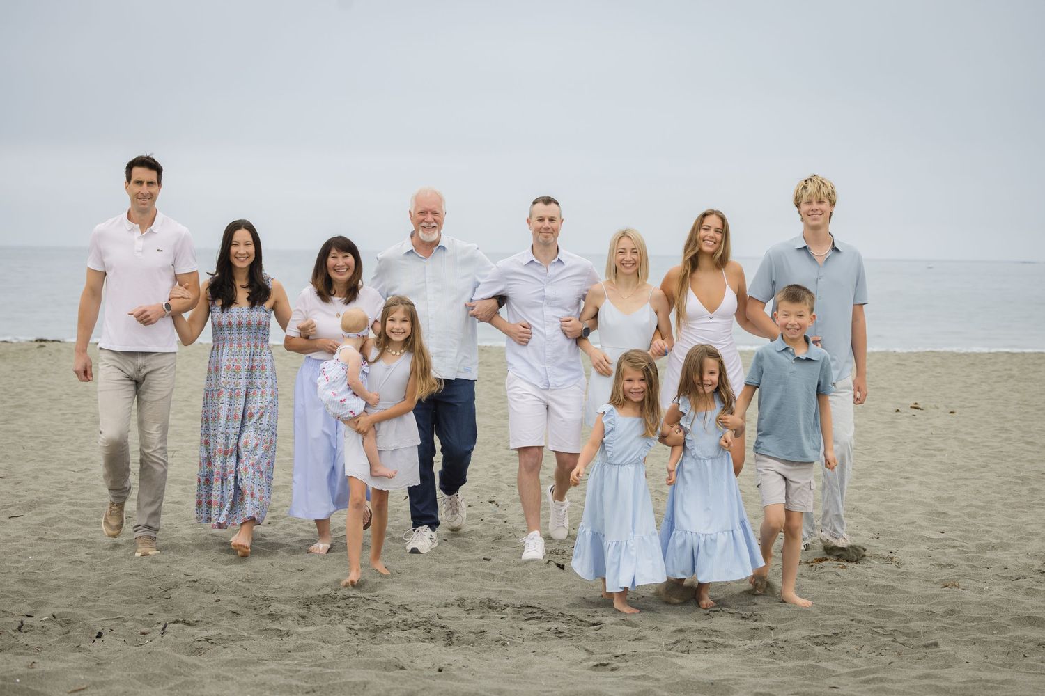 Large multigenerational family posing together on a sandy beach, wearing coordinated light blue and white outfits.