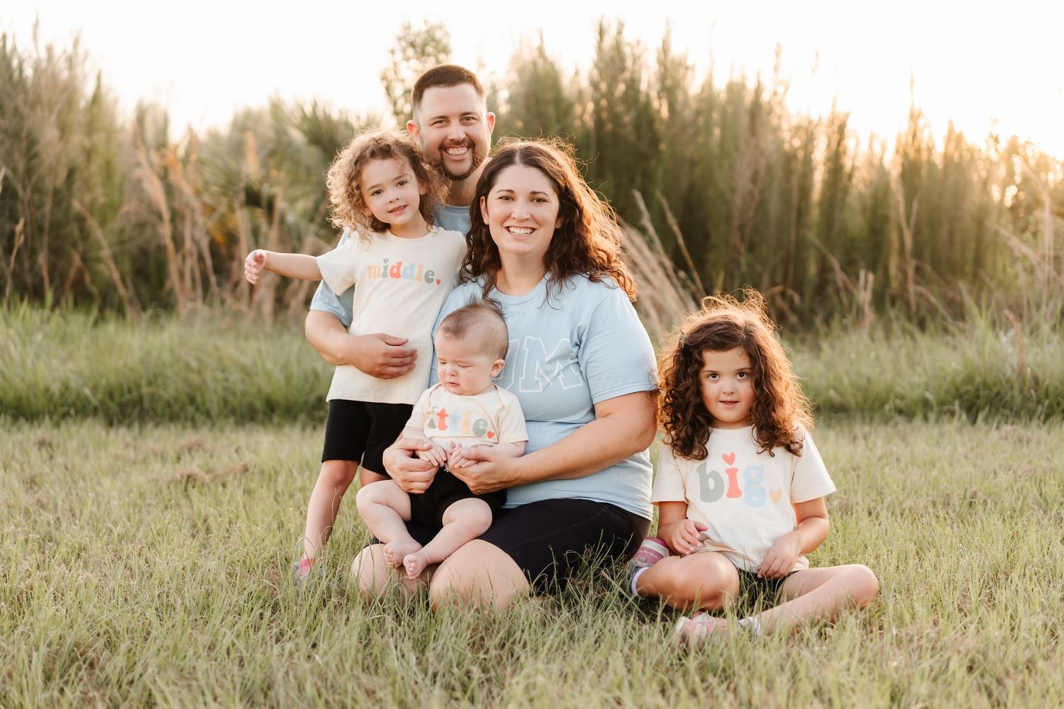 Family of five posing outdoors in a grassy field at golden hour, parents with three young children wearing matching white shirts.