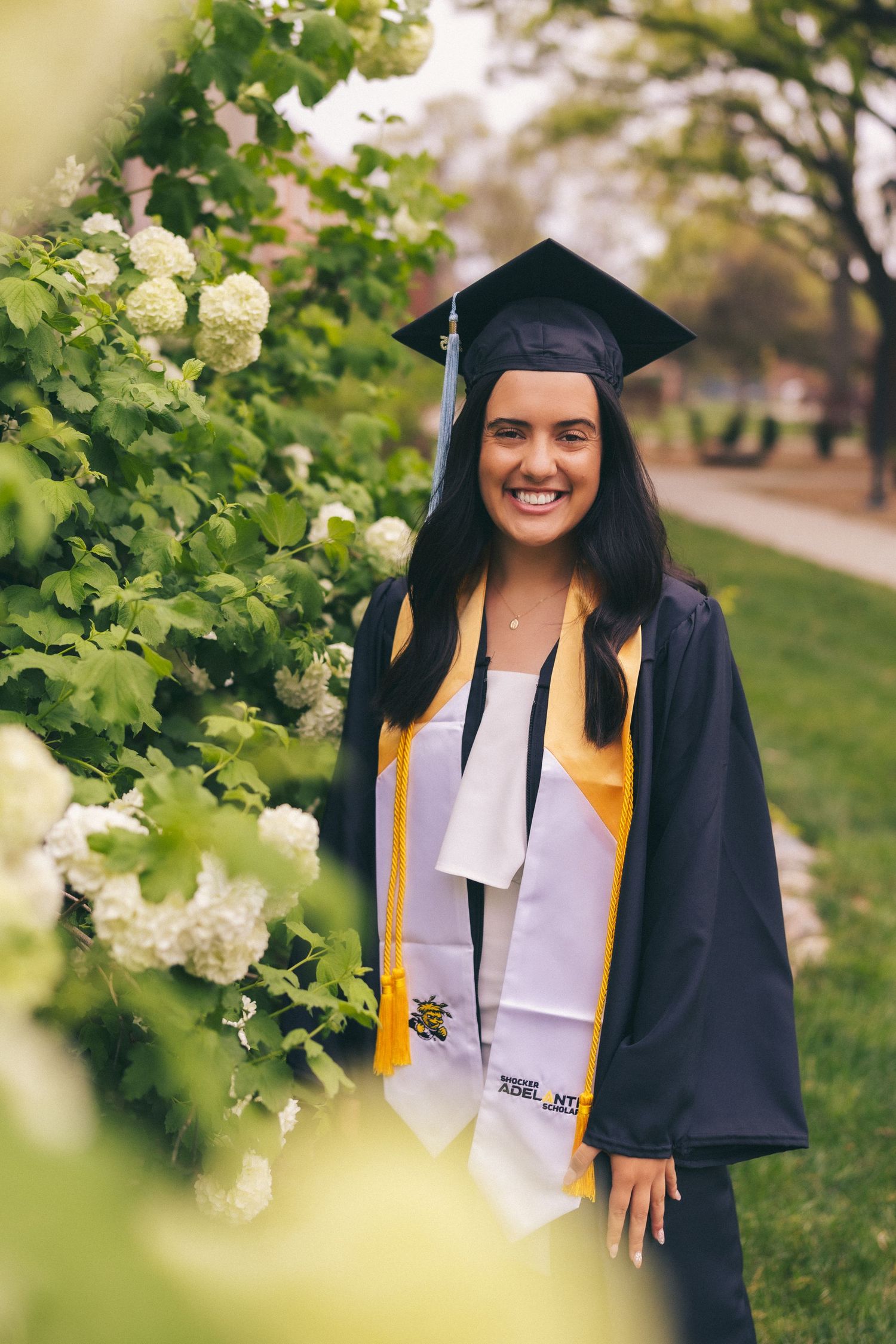 Graduate in cap and gown poses near blooming white flowers on a spring day.