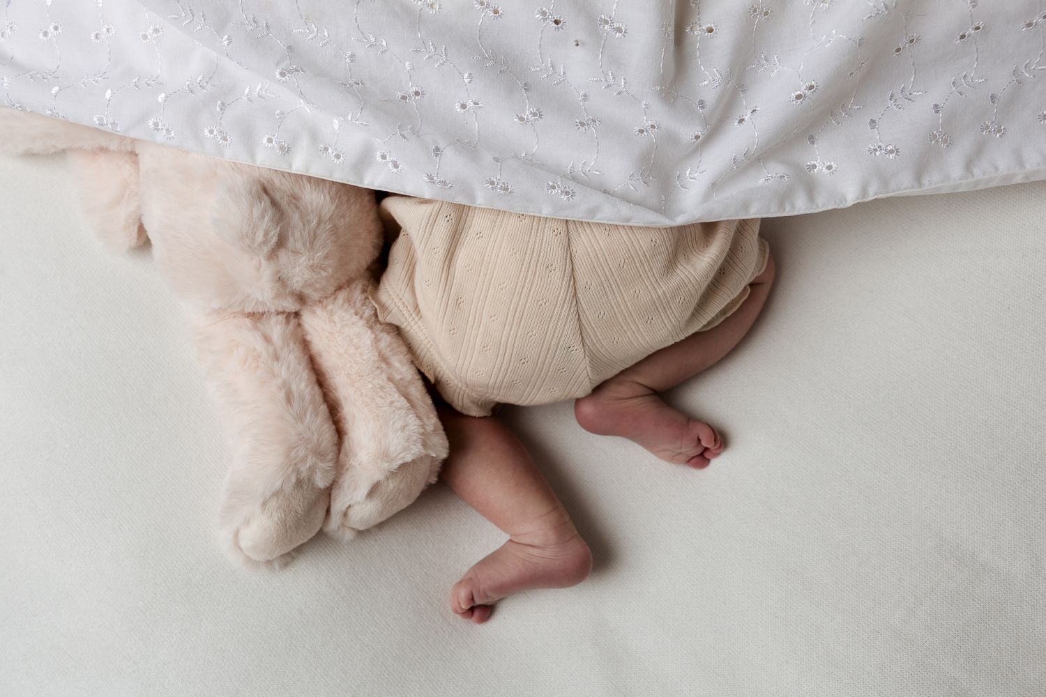 Baby sleeping under white blanket with a plush teddy bear, tiny feet peeking out on a soft white surface.