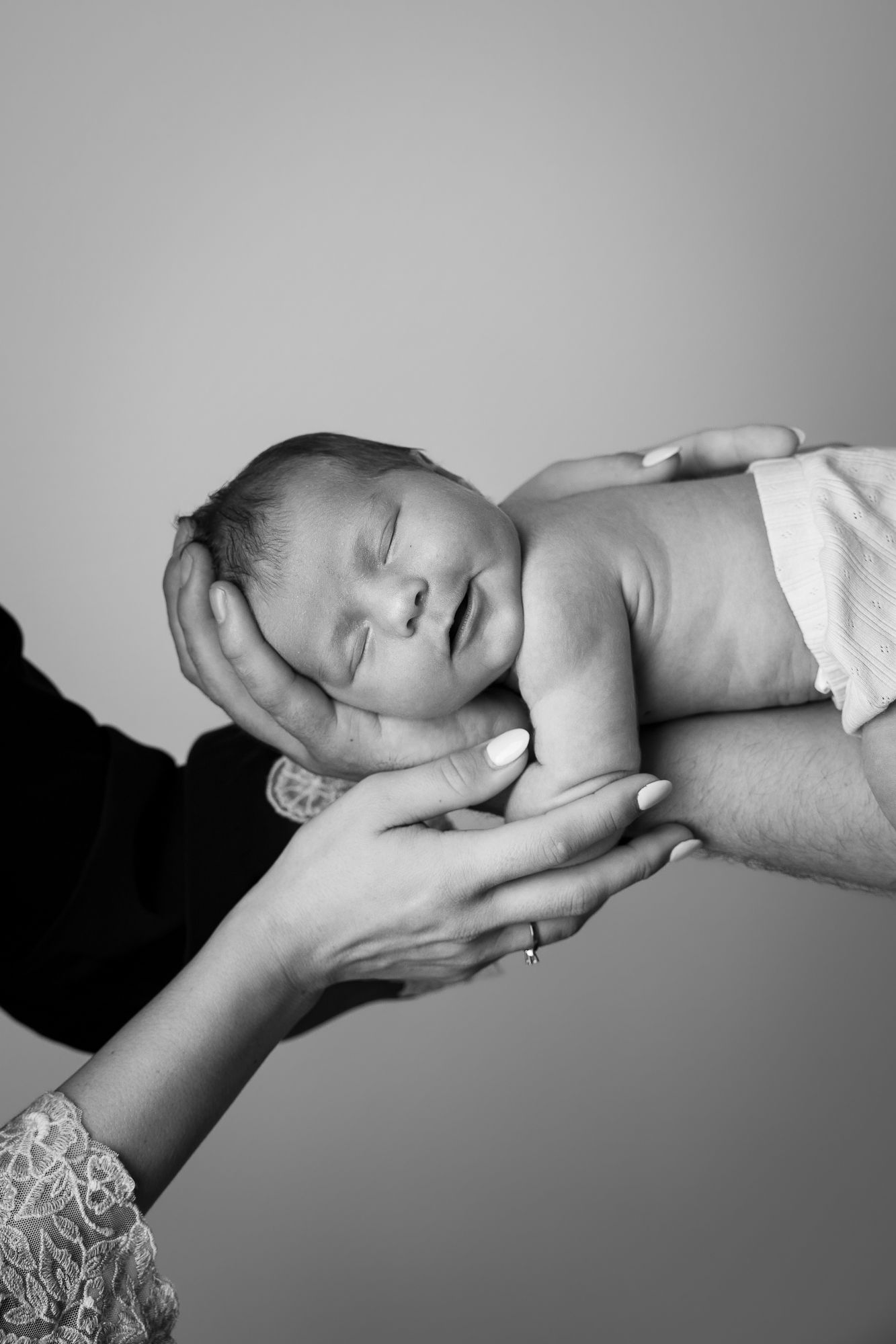 Black and white photo of a sleeping newborn baby cradled gently in the hands of two parents.