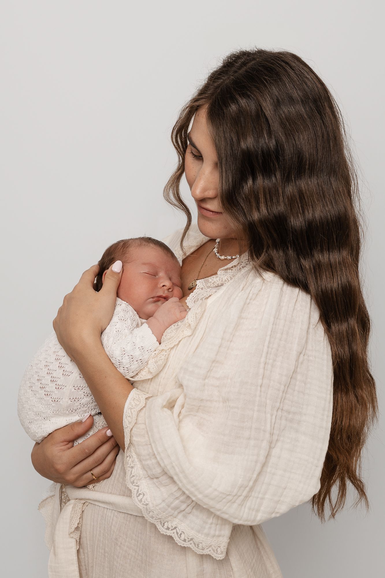 A young mother with long brown hair tenderly holds her sleeping newborn baby, both dressed in soft cream tones.