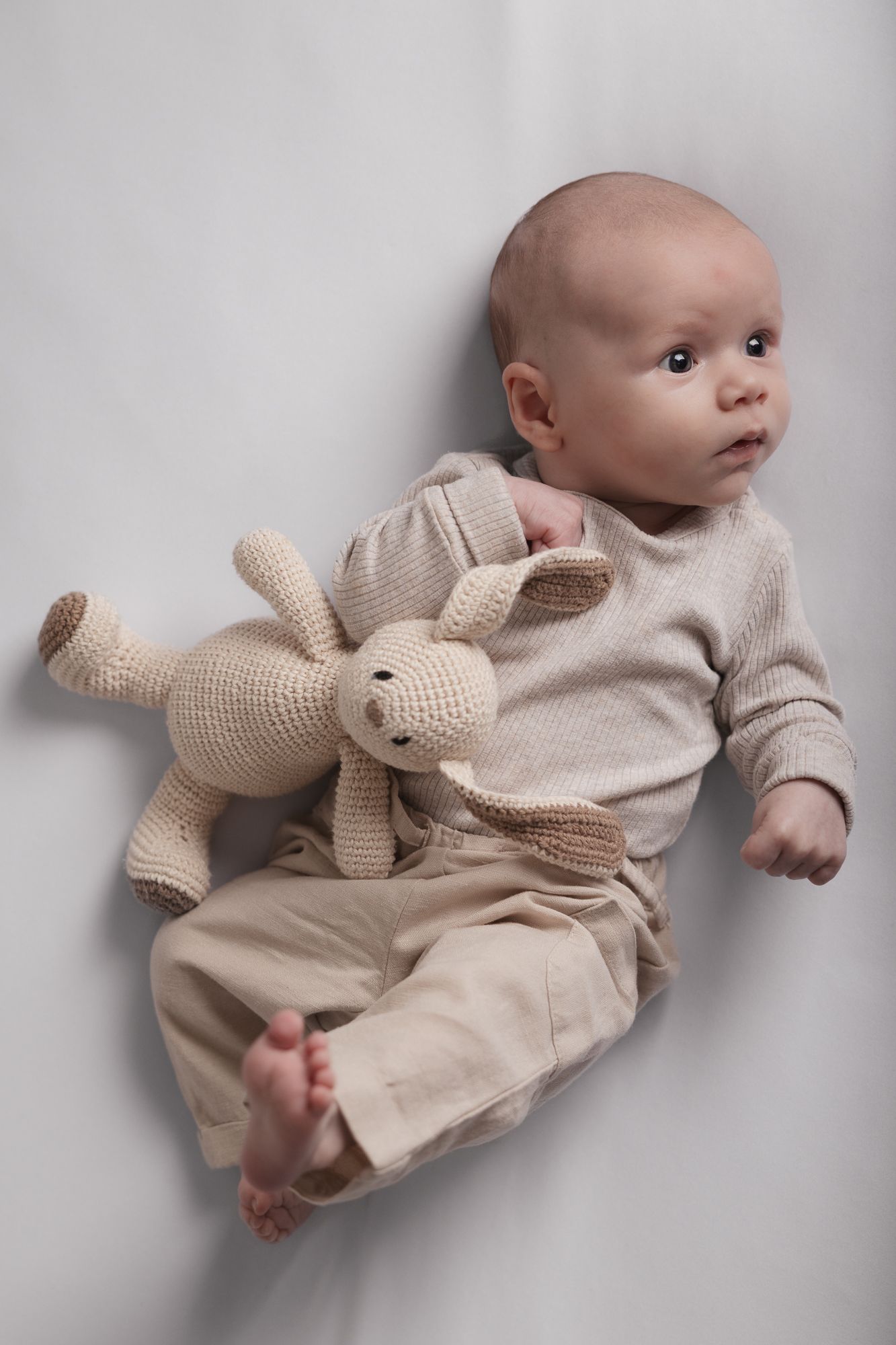 A newborn baby in beige knit outfit lying on white background holding a crocheted bunny toy.