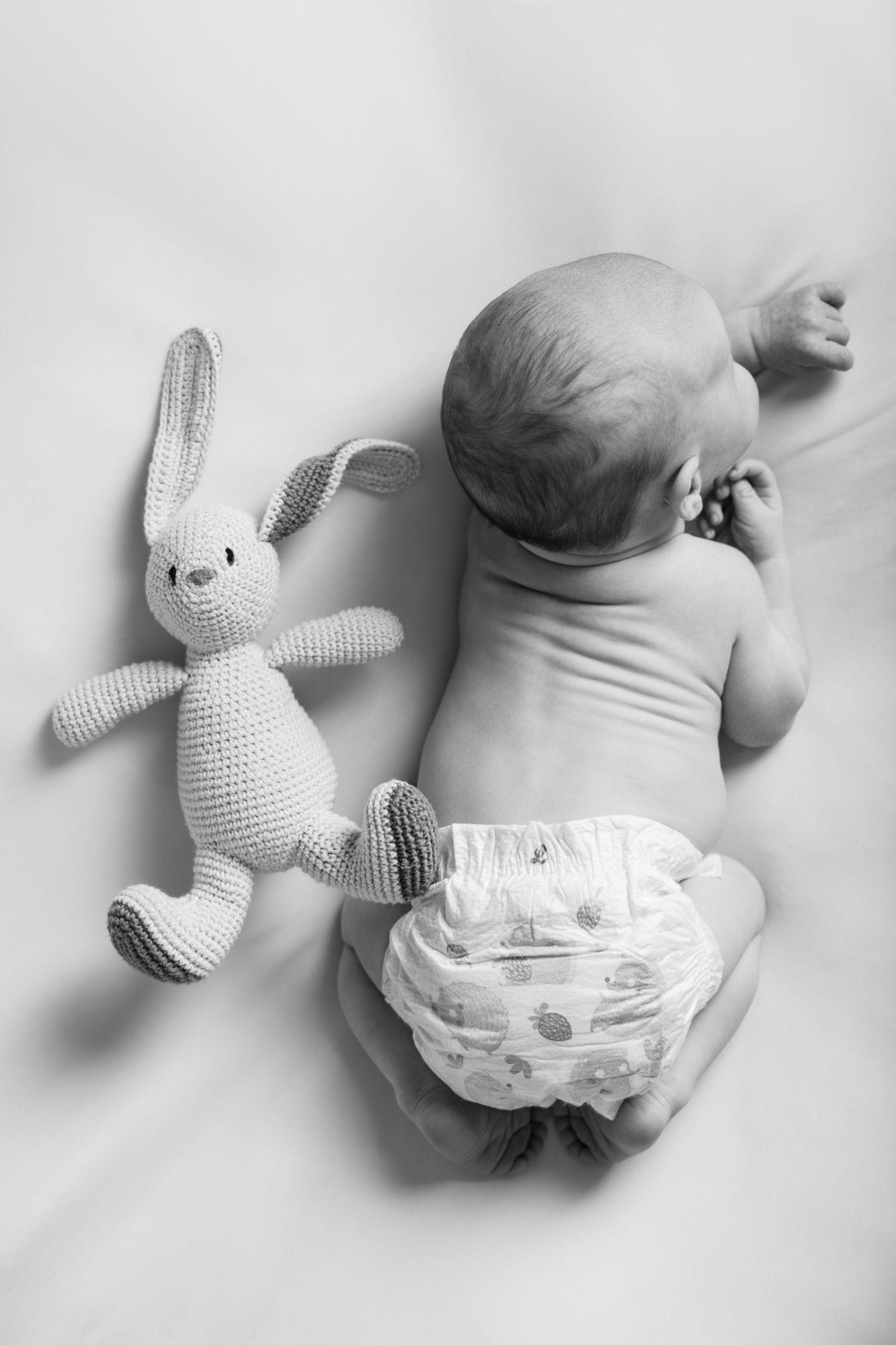 Black and white photo of a newborn baby lying on their stomach in a diaper beside a crocheted bunny toy.