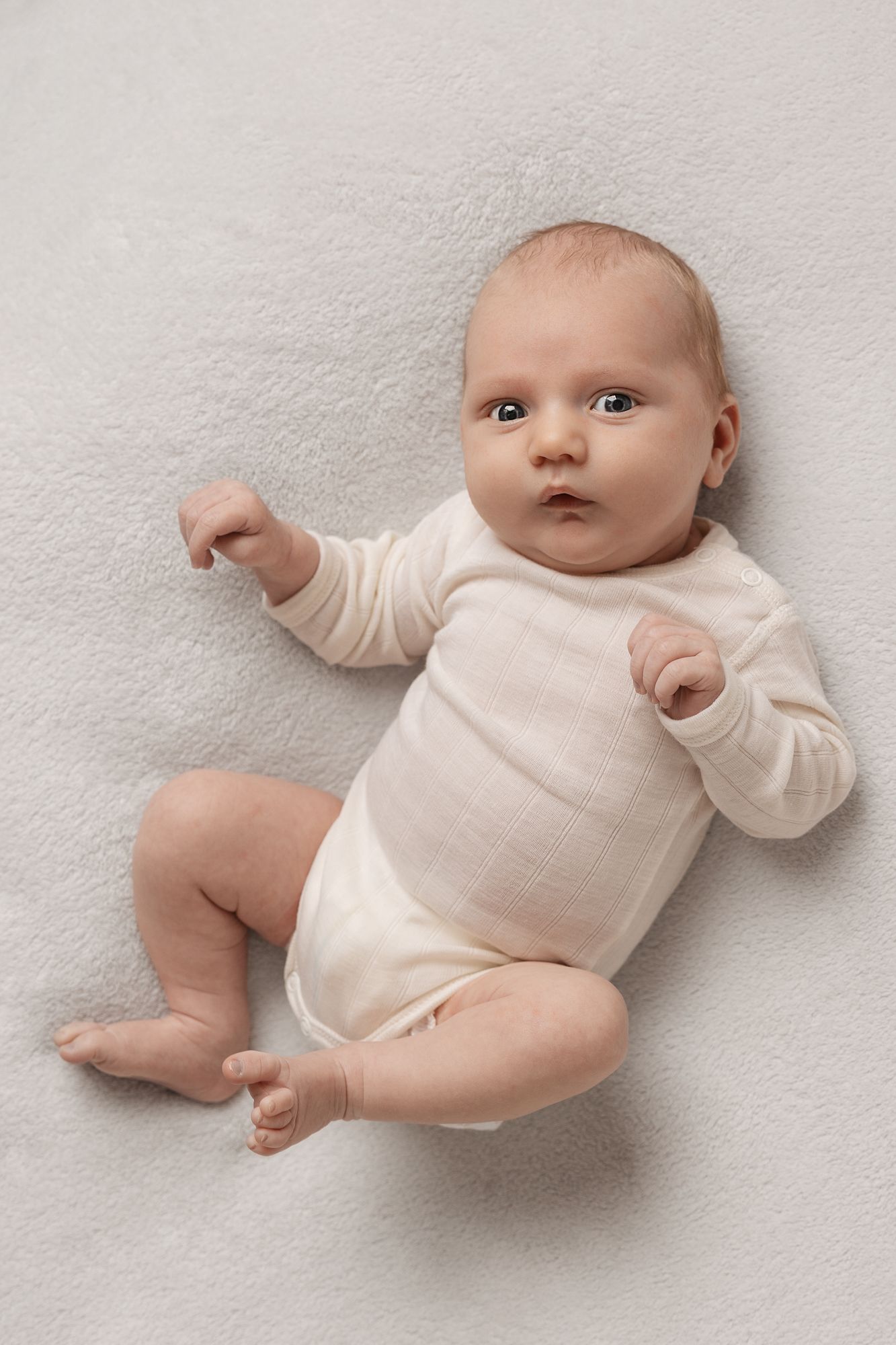 A cute baby in a white onesie lying on a soft white blanket, looking up with wide curious eyes.