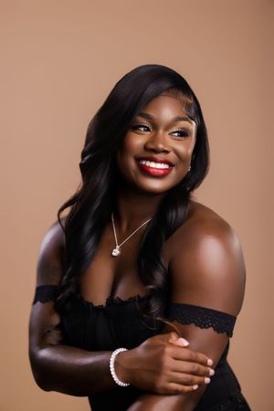 Smiling Black woman with long black wavy hair, red lips, pearl bracelet, and off-shoulder black top on beige backdrop.