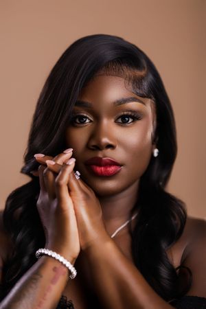 Black woman with long wavy black hair, bold red lips, pearl bracelet, and tattoo posing on a beige background.