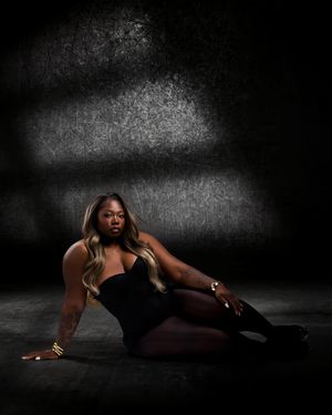 A woman in a black bodysuit and thigh-high boots poses dramatically on a dark studio floor under dramatic lighting.