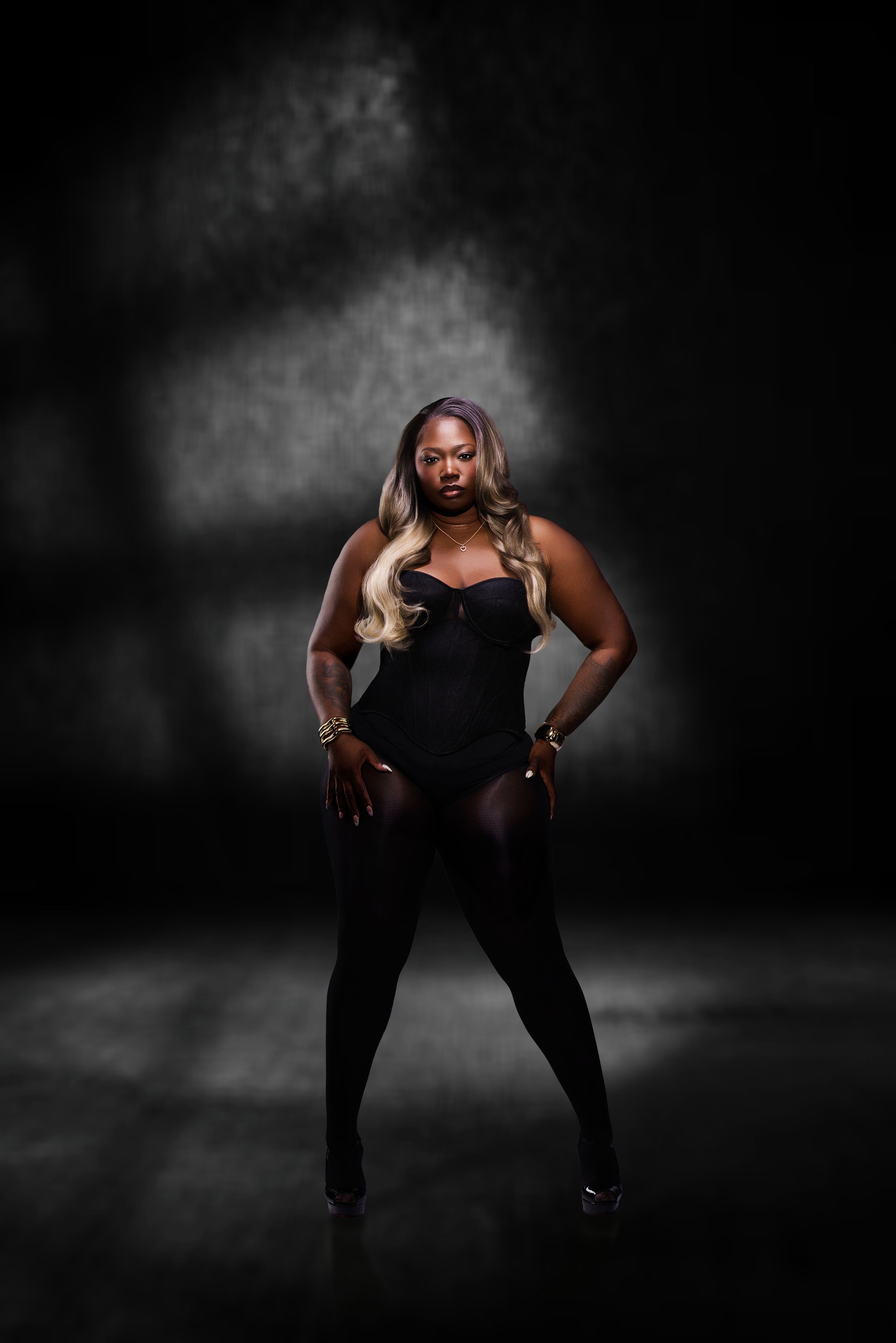 A confident woman in a black bodysuit and tights poses dramatically against a dark smoky studio background.