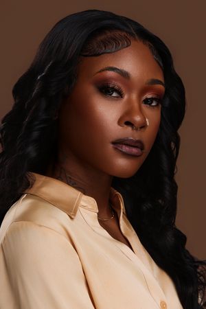 Black woman with long wavy black hair, warm smoky eye makeup, nose ring, and beige satin shirt on brown background.