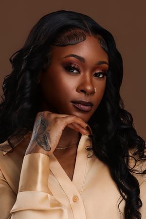 A Black woman with long wavy hair poses confidently in a gold satin blouse with a hand tattoo visible.