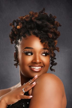 Smiling Black woman with decorative rhinestone eyebrows, auburn curly updo locs, rings, and bare shoulders against gray background.