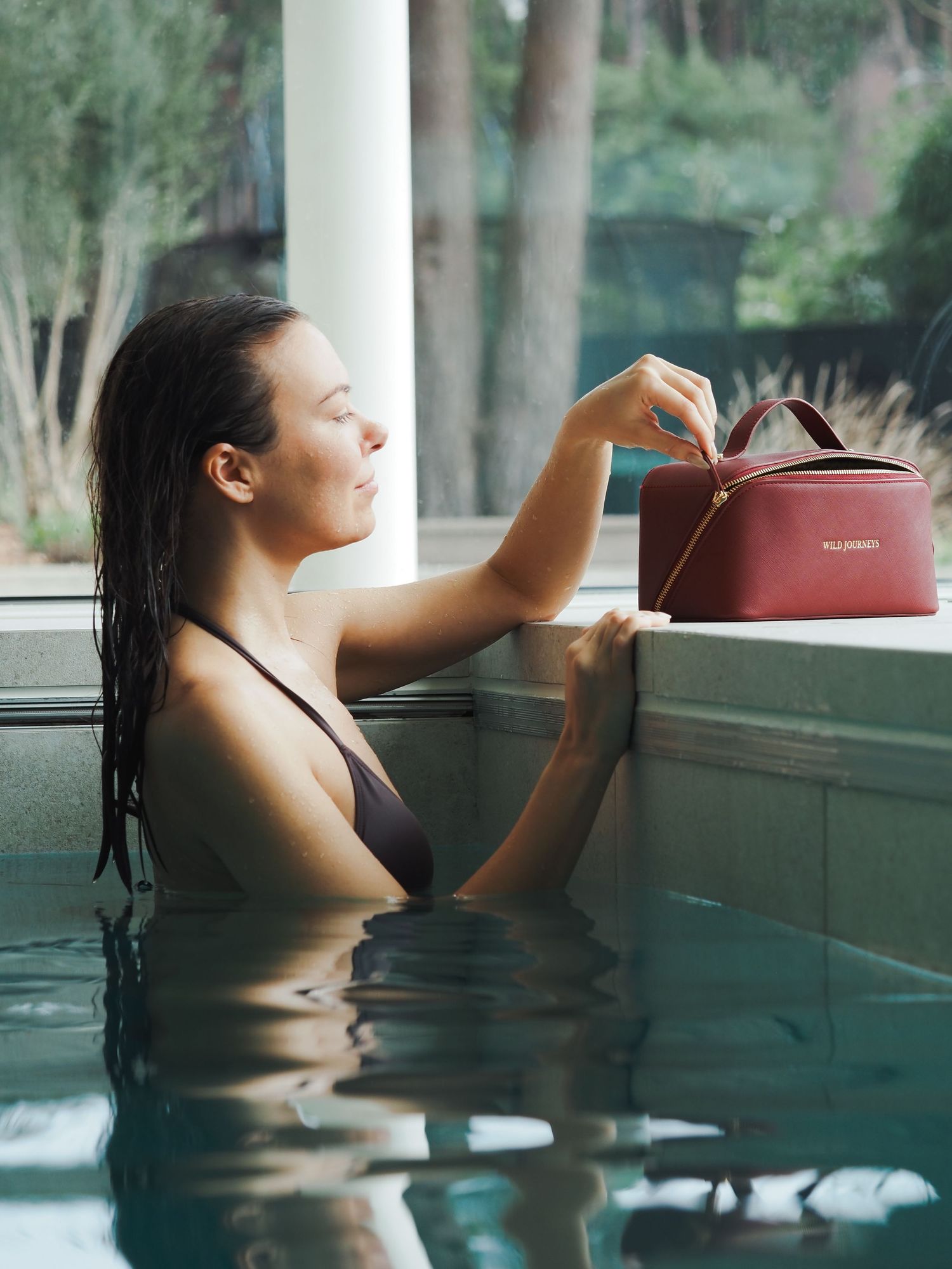 Woman in pool holding a red luxury handbag on pool edge in elegant indoor spa setting.
