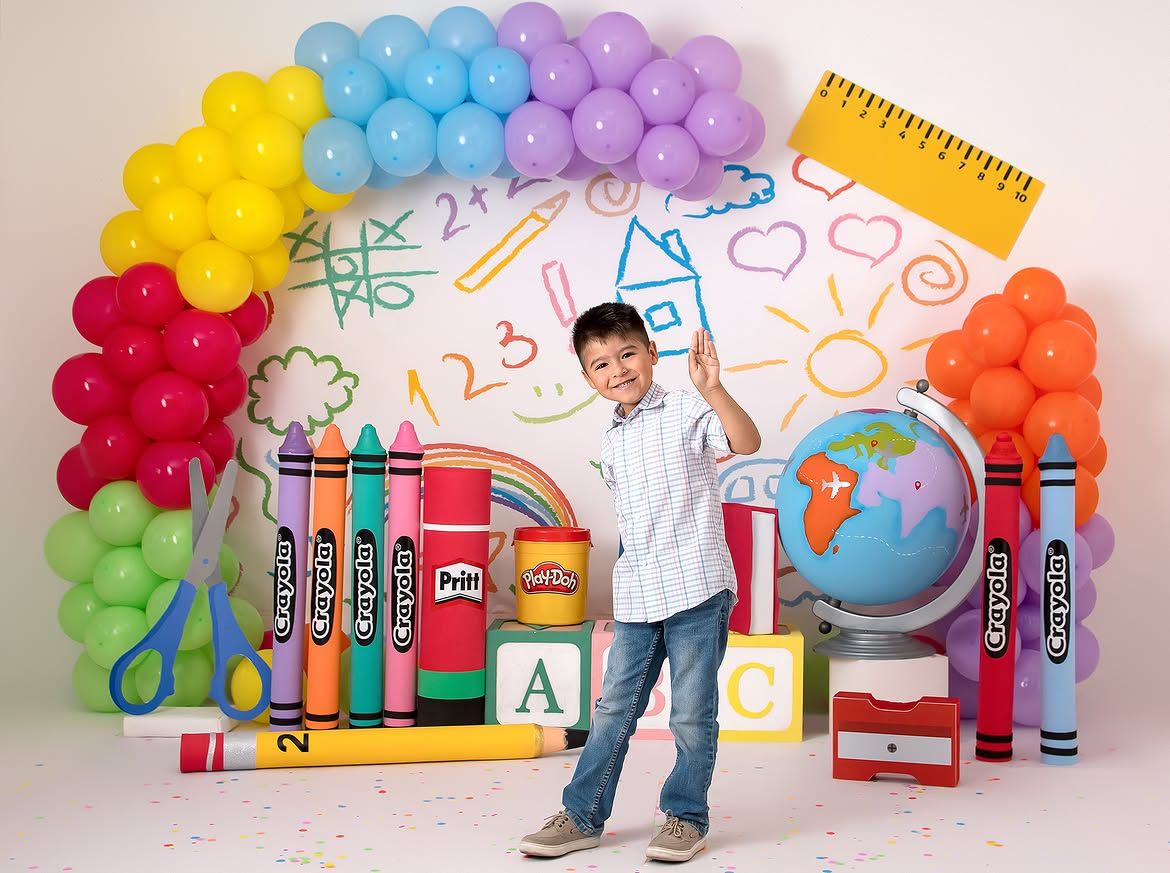 Boy posing in front of colorful back-to-school backdrop with balloons, crayons, globe, and ABC blocks.