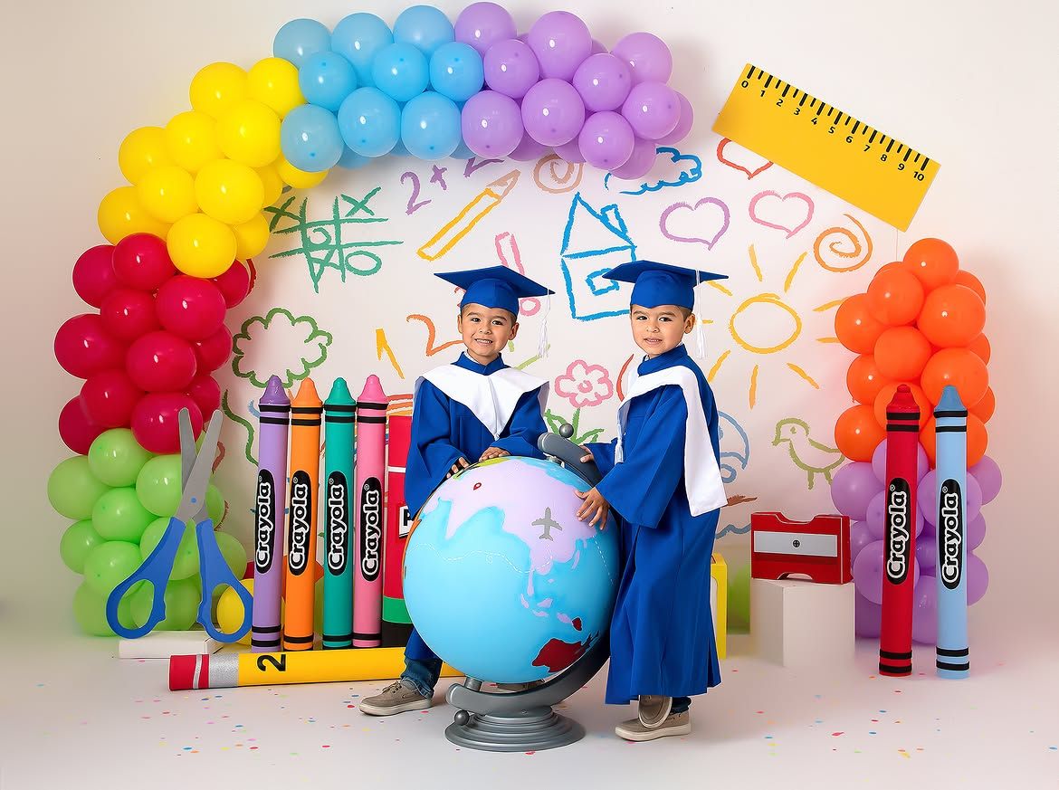 Two children in blue graduation caps and gowns pose with a globe amid colorful balloons and oversized school supply decorations.