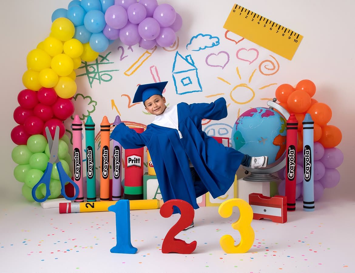 Child in blue graduation cap and gown poses with giant crayons, numbers 1-2-3, and colorful balloons backdrop.