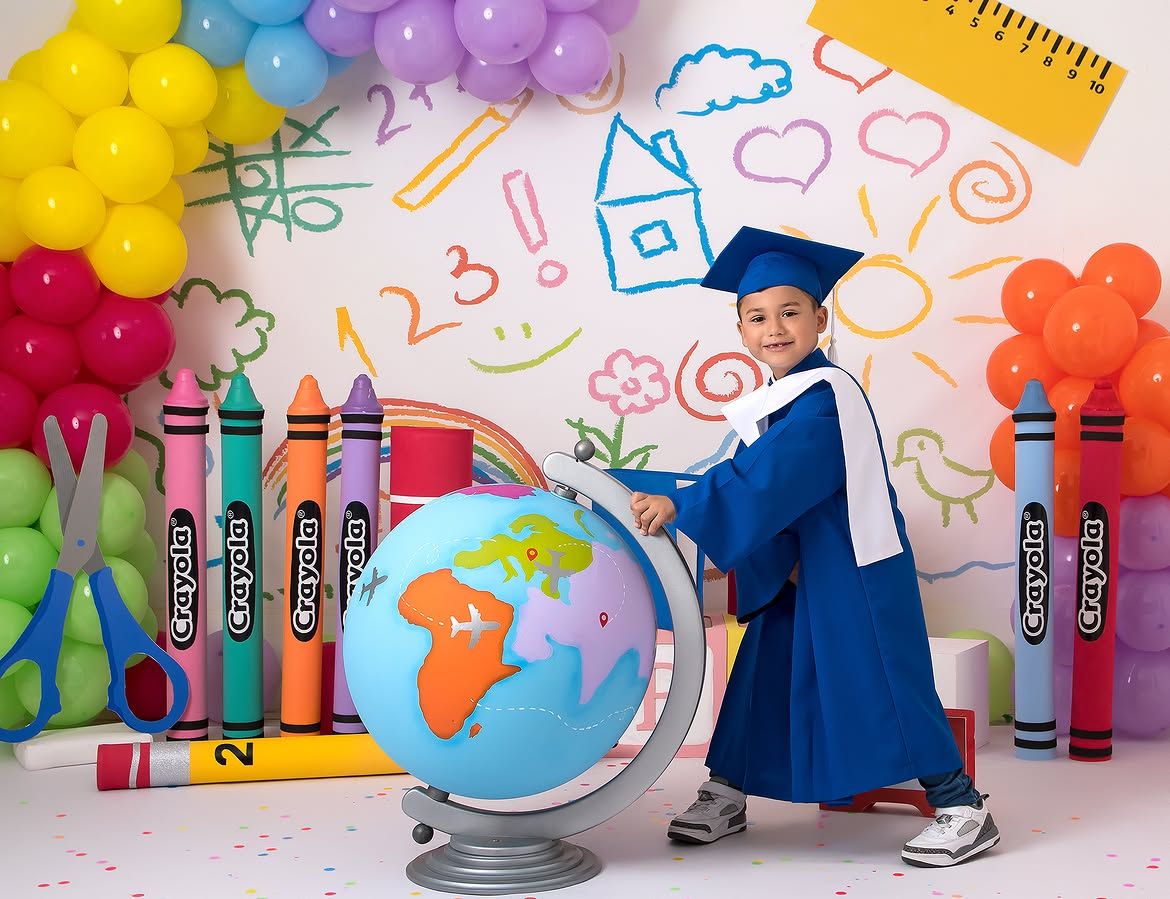 Young child in blue graduation cap and gown posing with a globe, surrounded by colorful balloons and Crayola crayons.