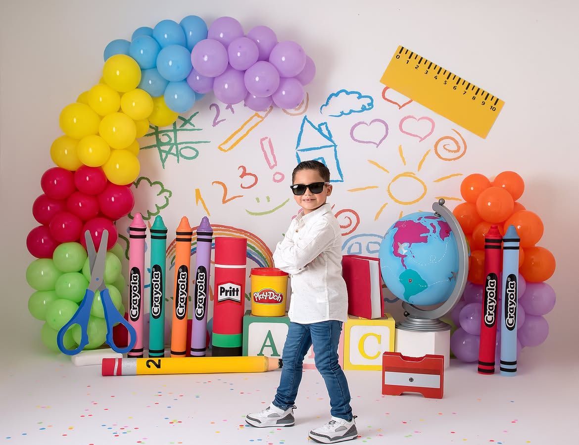 Child posing confidently in sunglasses among colorful school-themed balloons, giant crayons, and Play-Doh backdrop.