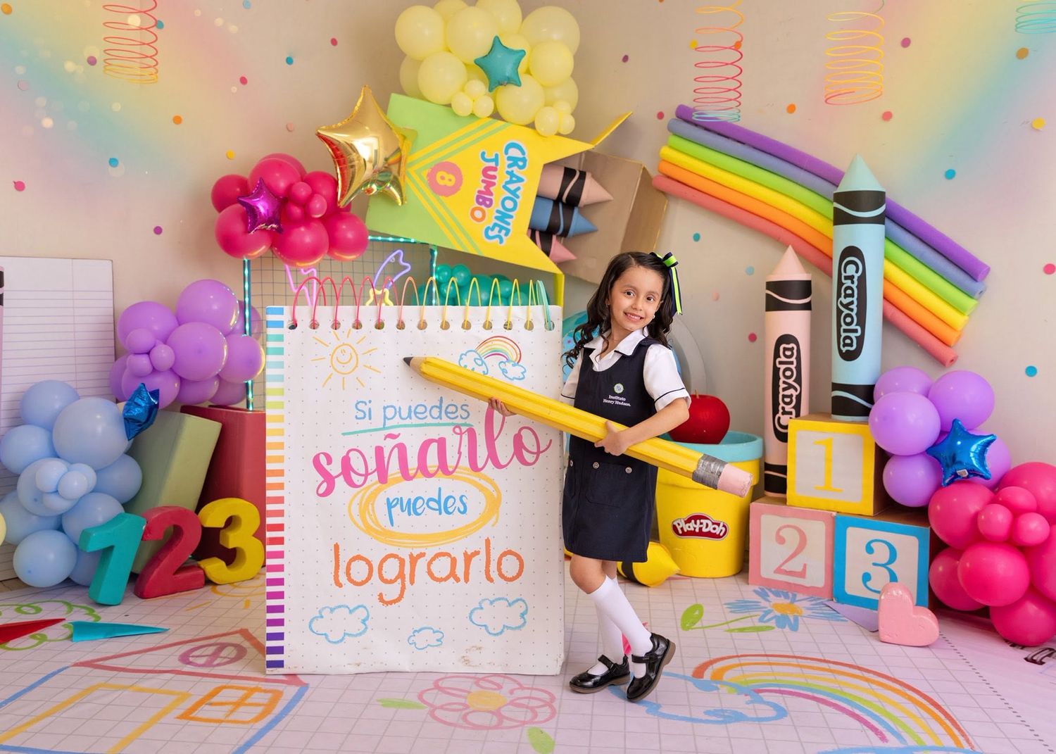 A young girl in school uniform poses by a colorful rainbow backdrop with balloons, blocks, and an inspirational Spanish quote on a notebook.