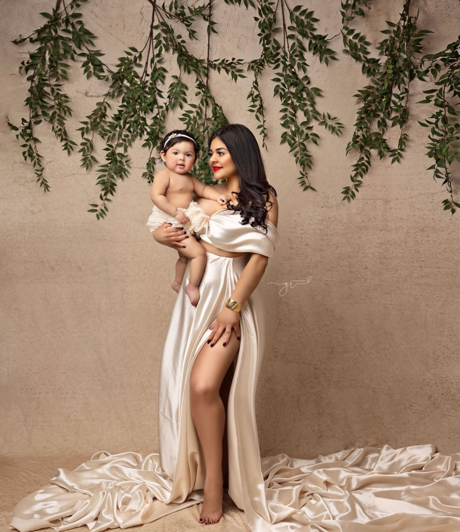 A woman in an elegant cream satin gown holds her baby girl in a studio with hanging greenery backdrop.