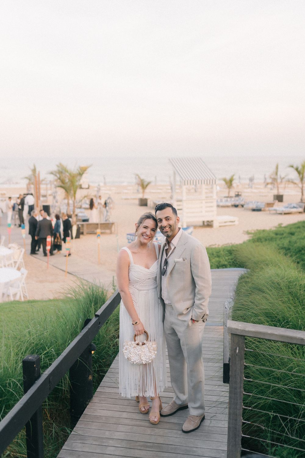 Couple in wedding attire smiling on a wooden boardwalk at a beachside venue during sunset.