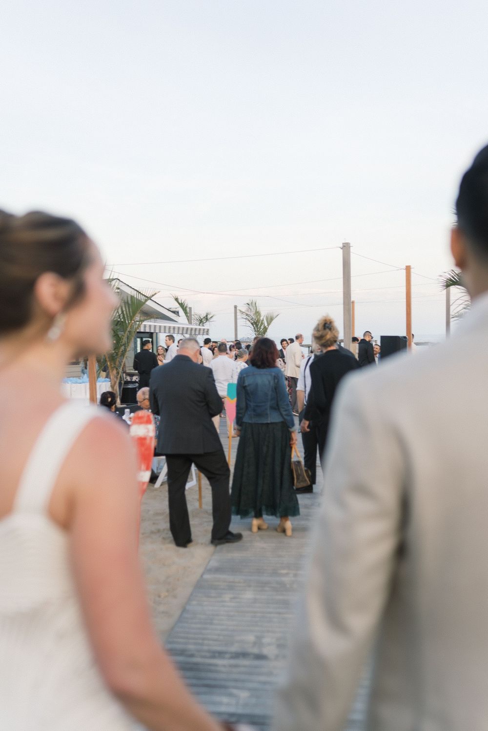 Guests gathered at an outdoor beach wedding reception on a sunny evening boardwalk.