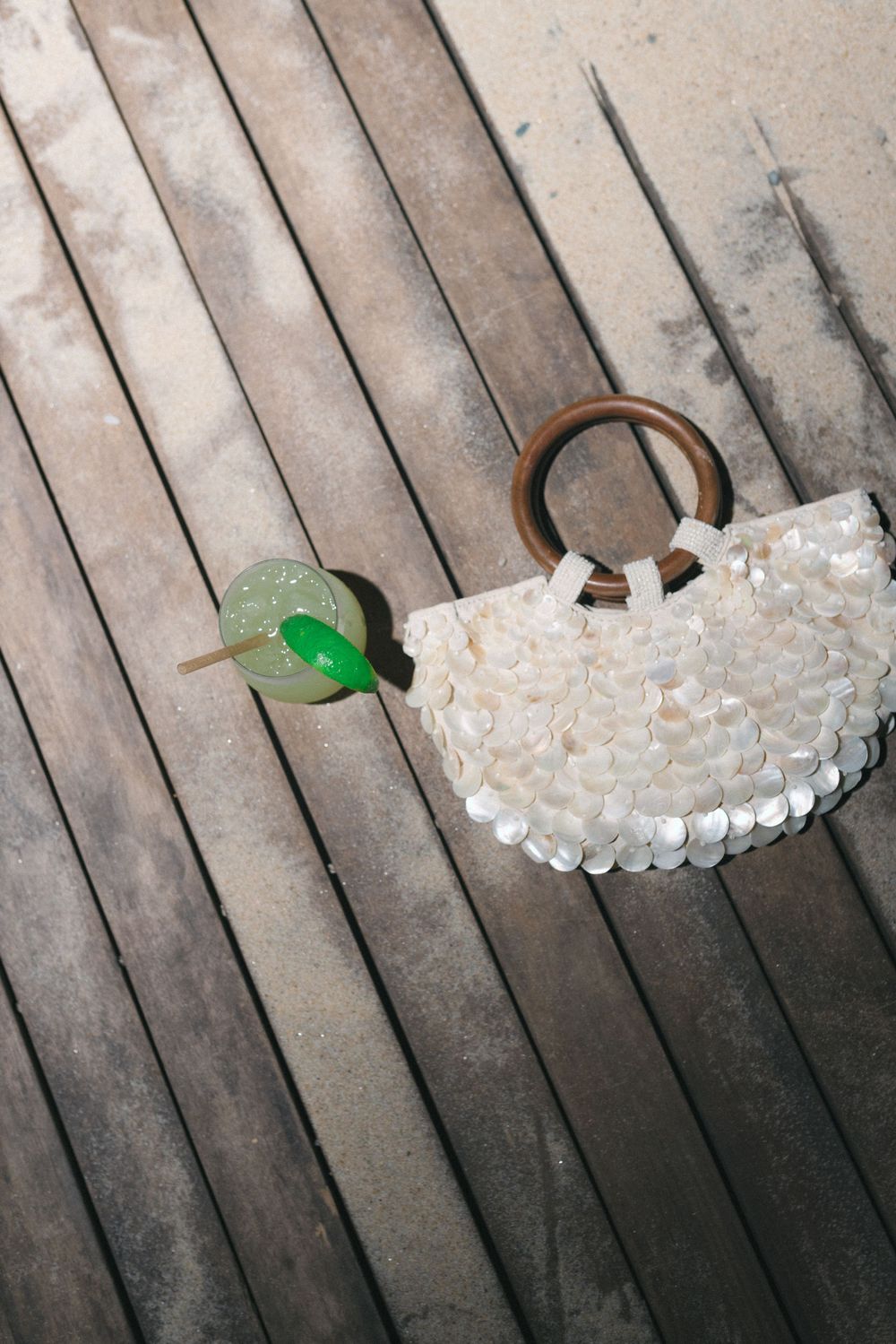 Shell purse with wooden handles beside a green cocktail on a rustic wooden deck.