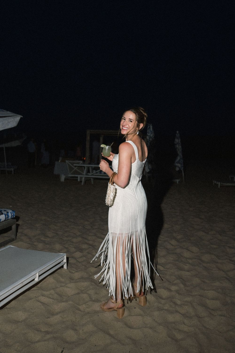 Woman in white fringe dress holding a drink and clutch bag at a nighttime beach wedding reception.