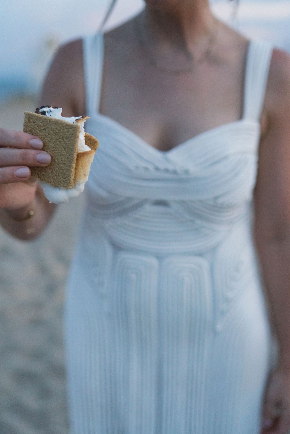 Woman in white dress holding a s'more on a sandy beach.