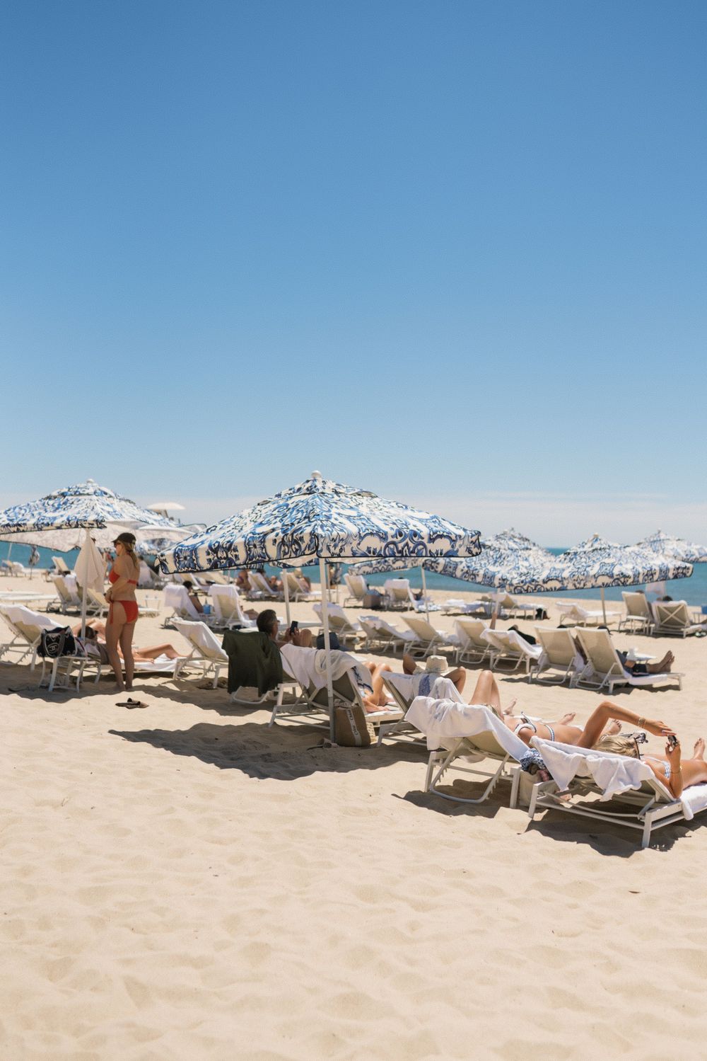 Beachgoers relaxing on lounge chairs under blue patterned umbrellas on a sunny sandy beach.