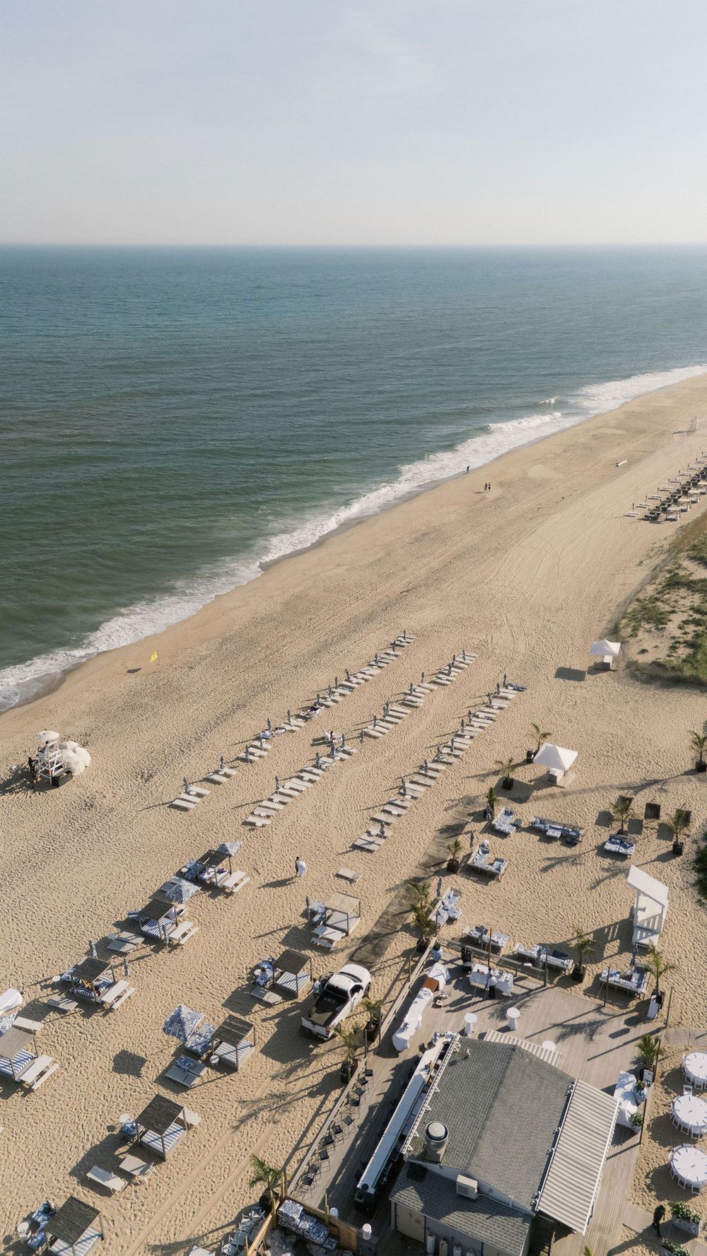 Aerial view of a pristine beach with rows of white lounge chairs, ocean waves, and a coastal venue below.