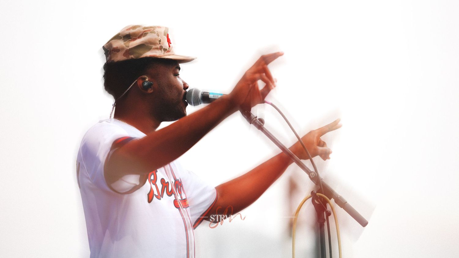 A Black male rapper performing on stage, pointing at the crowd while speaking into a microphone, wearing a white tee and cap.