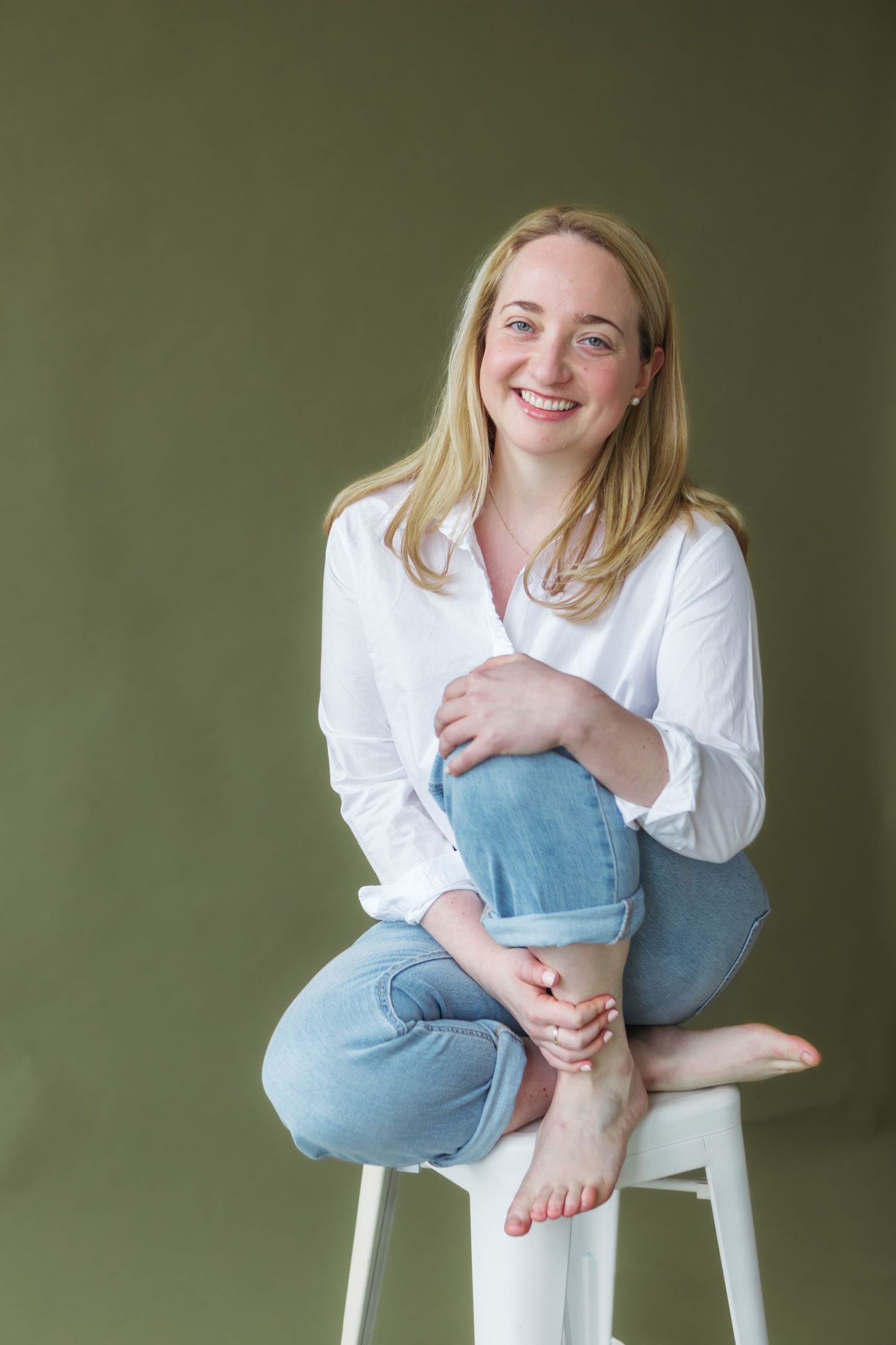 Smiling blonde woman in white shirt and jeans sitting cross-legged on white stool against olive green background.