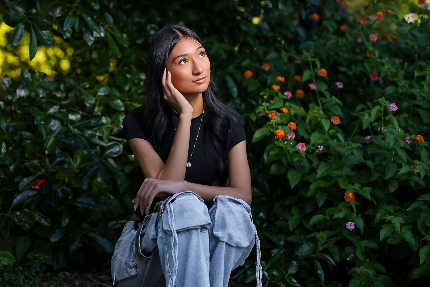Young woman with long dark hair sits pensively among vibrant garden flowers, wearing casual jeans and black top.