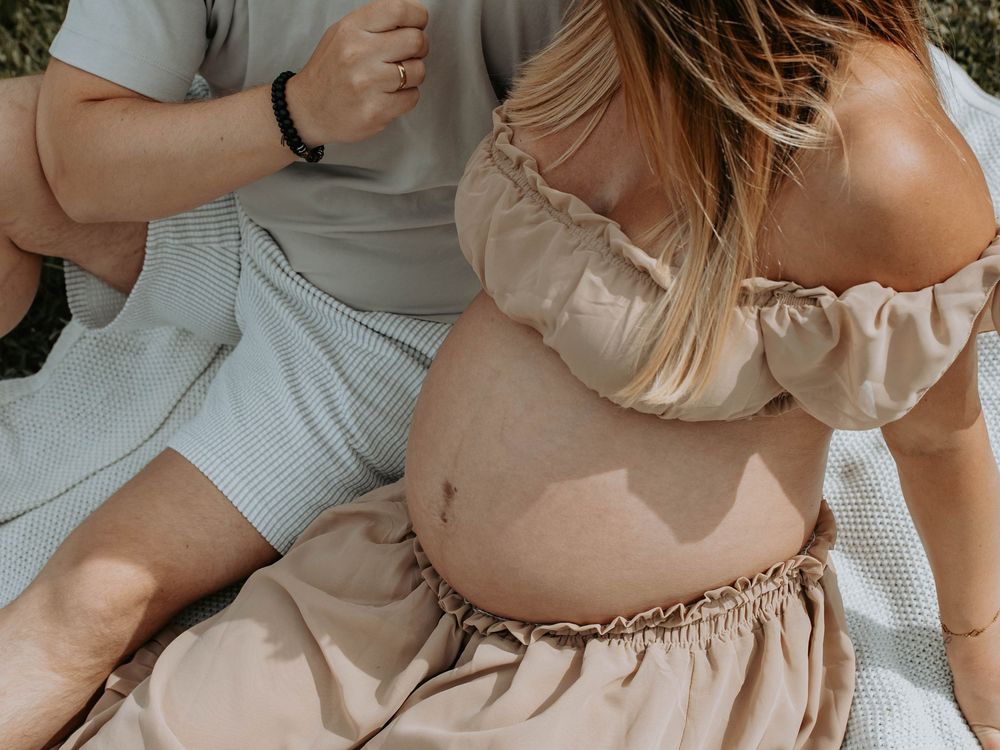 Seated pregnant woman in beige outfit with vitiligo on belly, partner beside her on blanket outdoors.