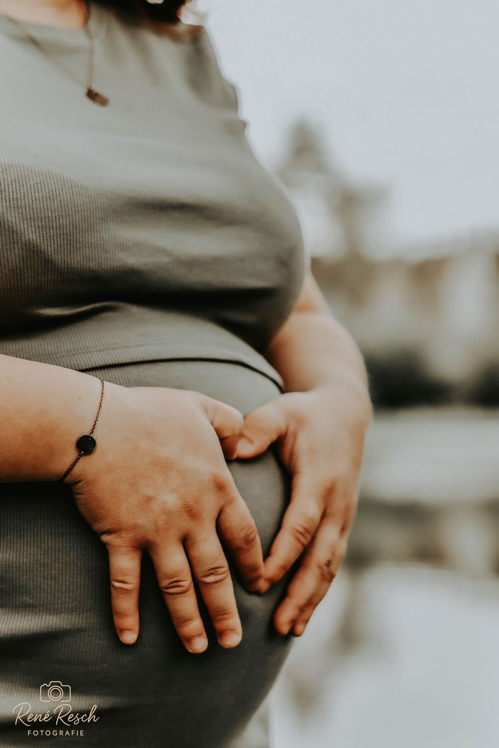 Close-up of pregnant woman cradling baby bump with both hands, wearing a sage green dress and delicate bracelet.