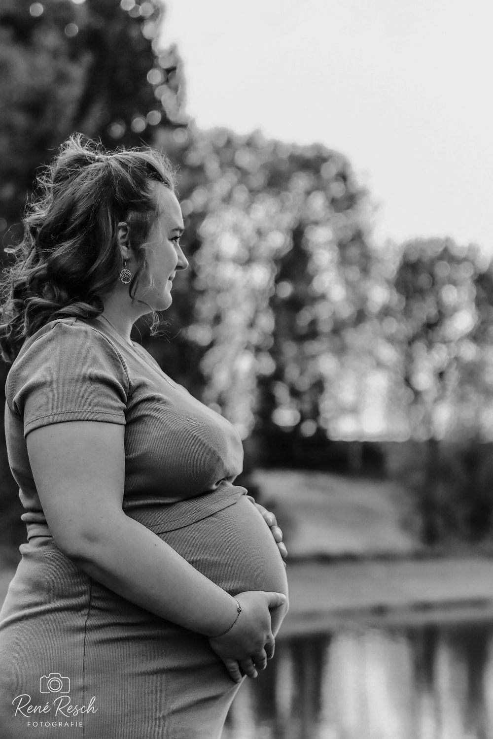 Black and white profile portrait of a pregnant woman in a fitted dress gently holding her baby bump outdoors.