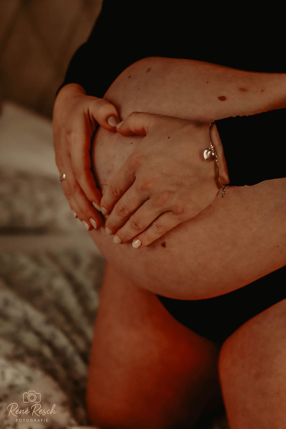 Pregnant woman cradling baby bump with both hands, wearing a bracelet, in a warm-toned maternity photography session.
