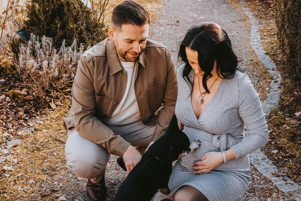 Couple crouching outdoors on a gravel path, man in brown jacket and pregnant woman in gray dress, sharing a tender moment.