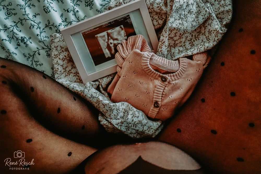 Flat lay of a pink knit baby sweater and framed photo on floral fabric, perfect for maternity announcement.