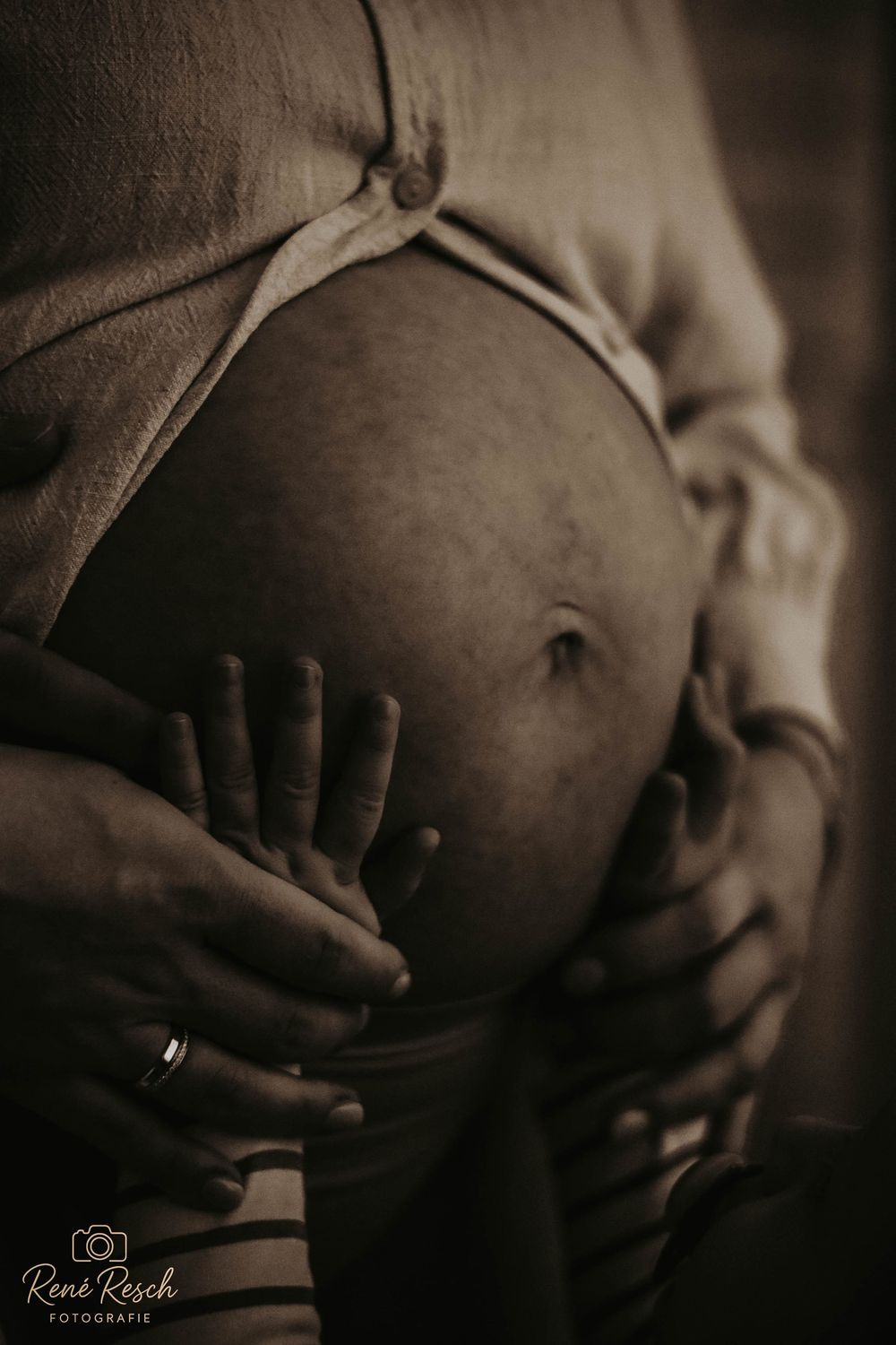 Black and white maternity photo of a pregnant belly being cradled by two pairs of hands, one wearing a wedding ring.
