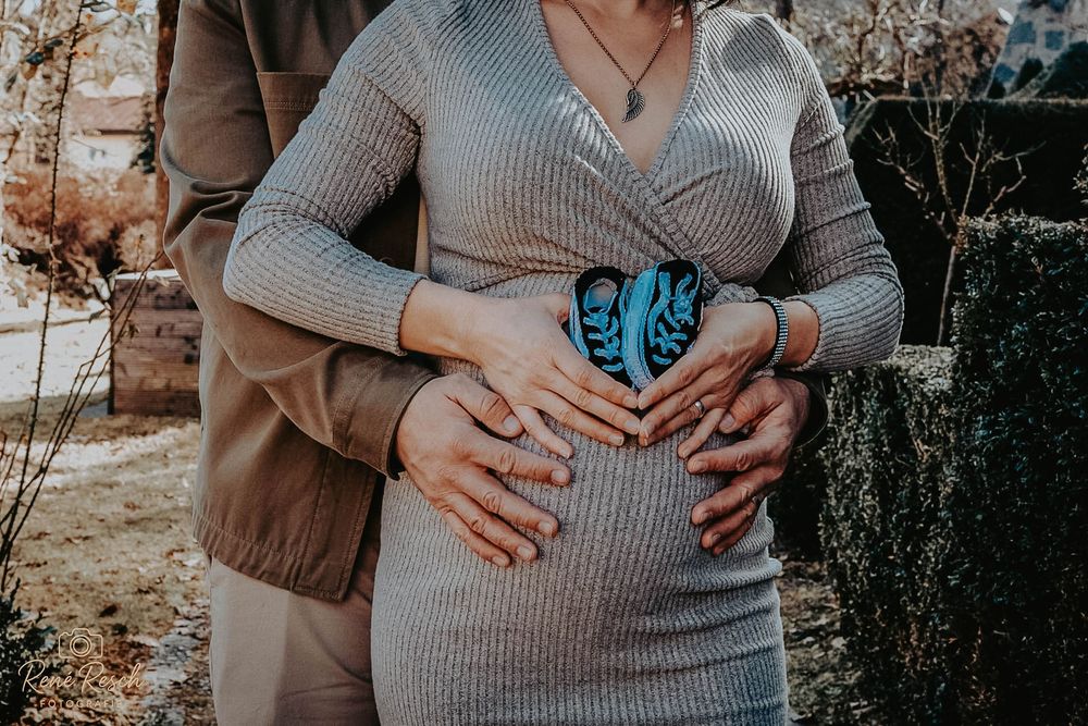 Pregnant woman in gray dress with partner's hands on baby bump outdoors.