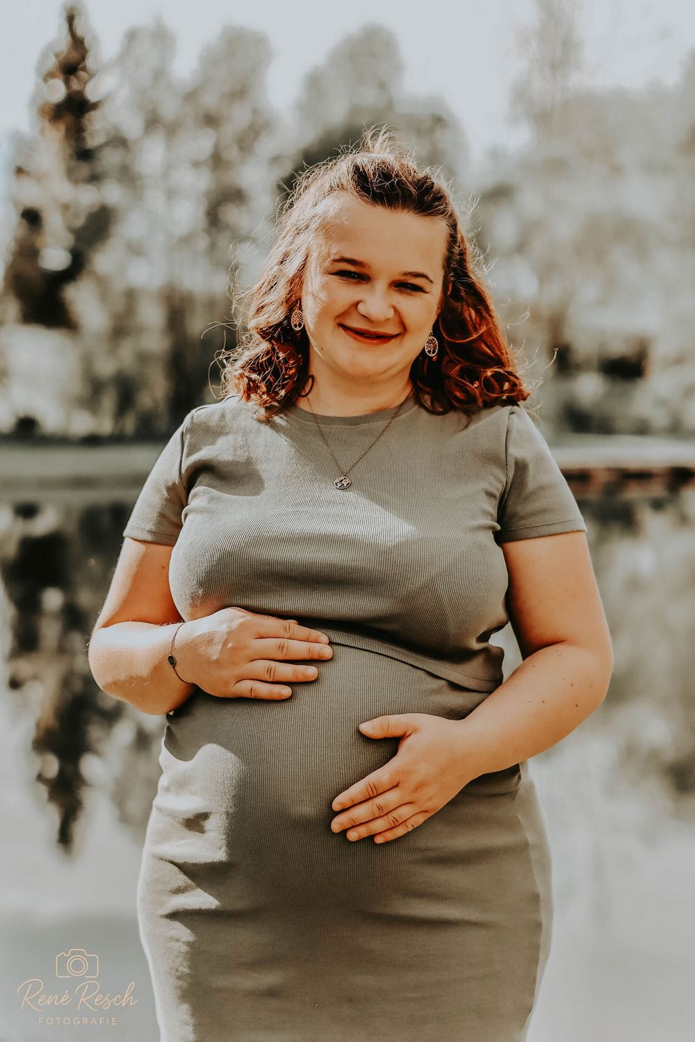Smiling pregnant woman in a gray fitted dress cradling her baby bump outdoors near a reflective pond.