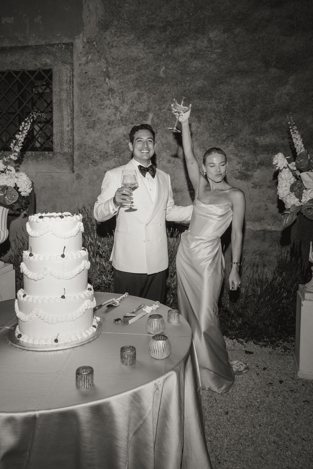 Newlyweds celebrating with champagne toast beside their white tiered wedding cake in black and white photo.