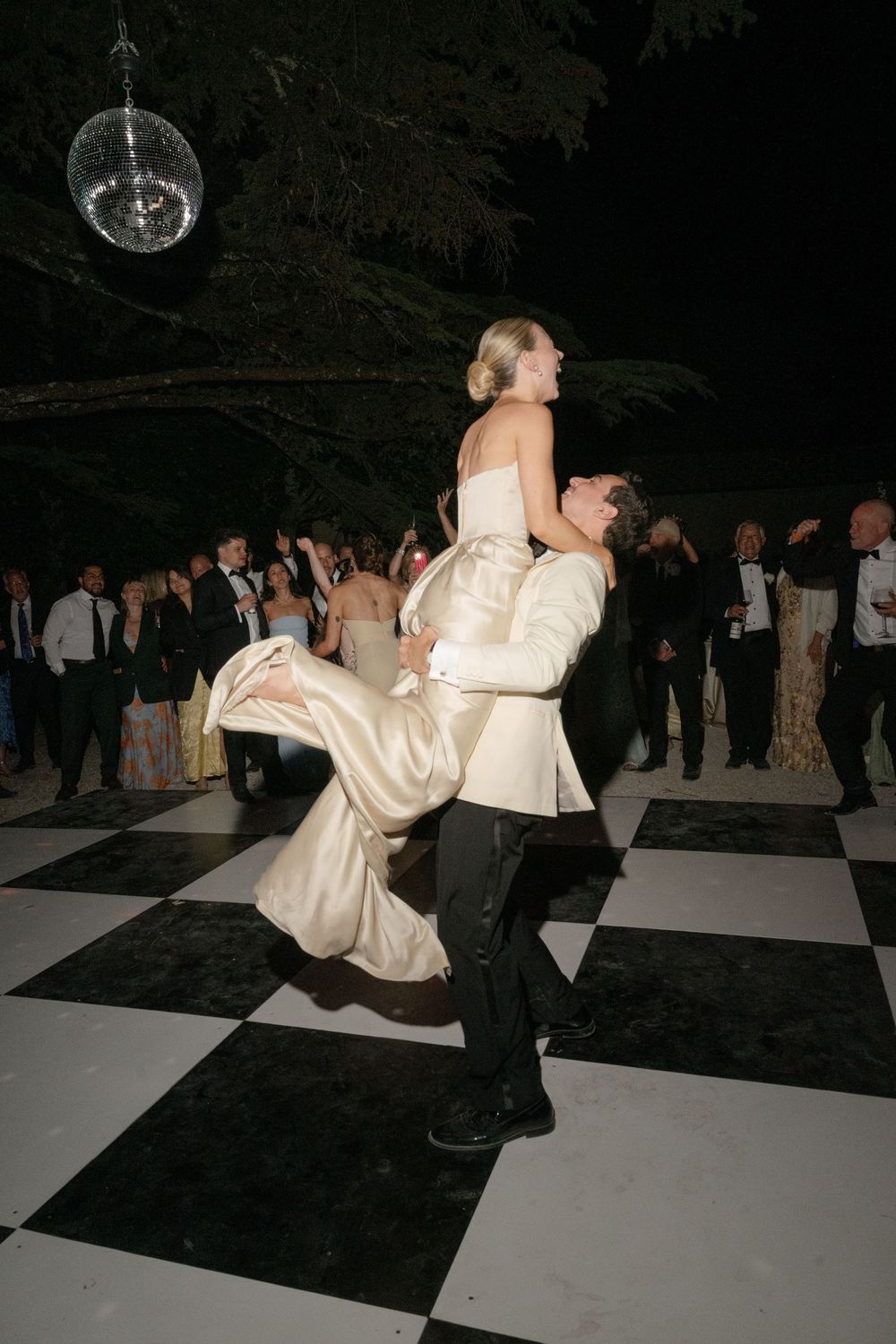 Groom lifts bride on black and white checkered dance floor at elegant outdoor wedding reception under disco ball.