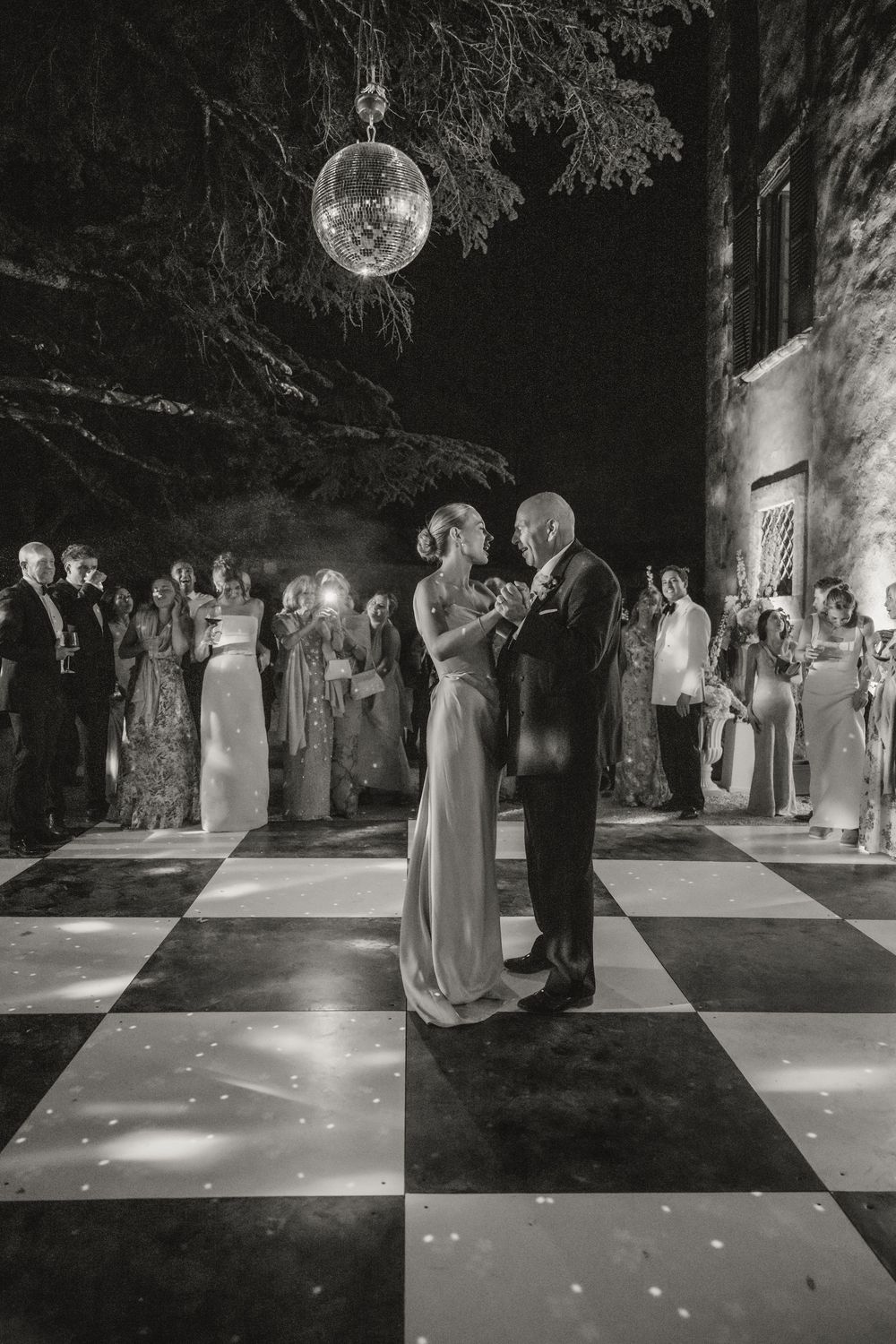 Couple shares first dance on checkered floor under disco ball at outdoor wedding reception with guests watching.